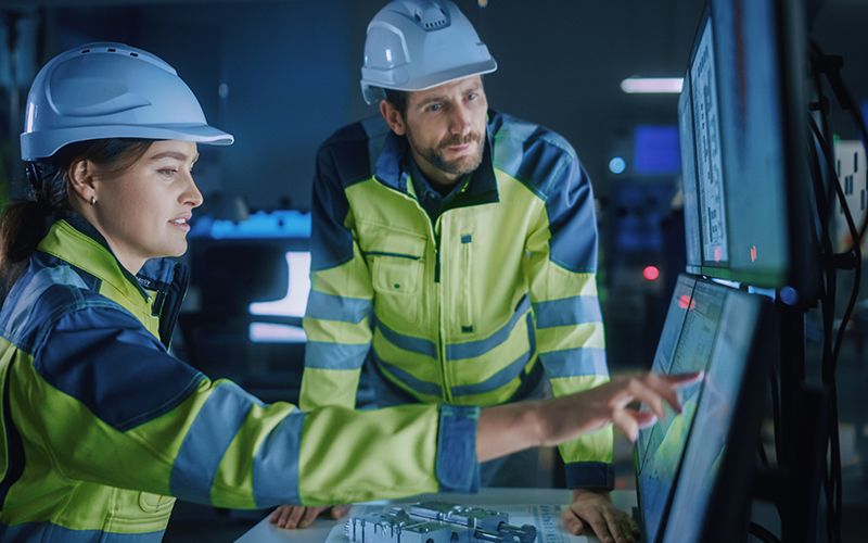 Two-engineers-working-with-data-wearing-safety-gear-one-pointing-at-screen